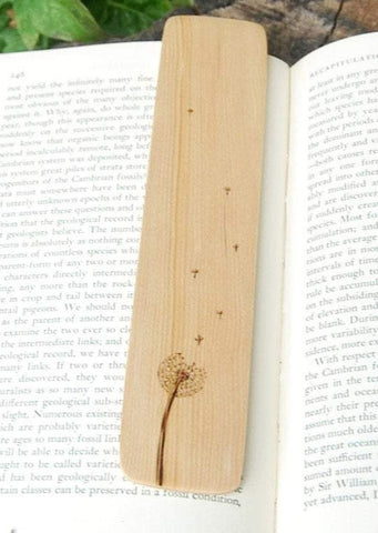 A photo of a bookmark laying on an open book with a leafy green background. On the bookmark is burned an image of a dandelion flower gone to seed, with some seeds floating free in the wind.