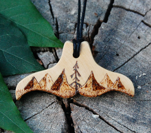 Whale tail shaped yellow cedar pendant on a wood background with leaves. The pendant has a mountain landscape with evergreen trees burned on to the face.
