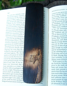 A photo of a woodburned bookmark resting on a book with a leafy background. The bookmark is burned mostly black, with a cloaked figure with a shadowed face holding a lantern that casts an eerie pool of light. 
